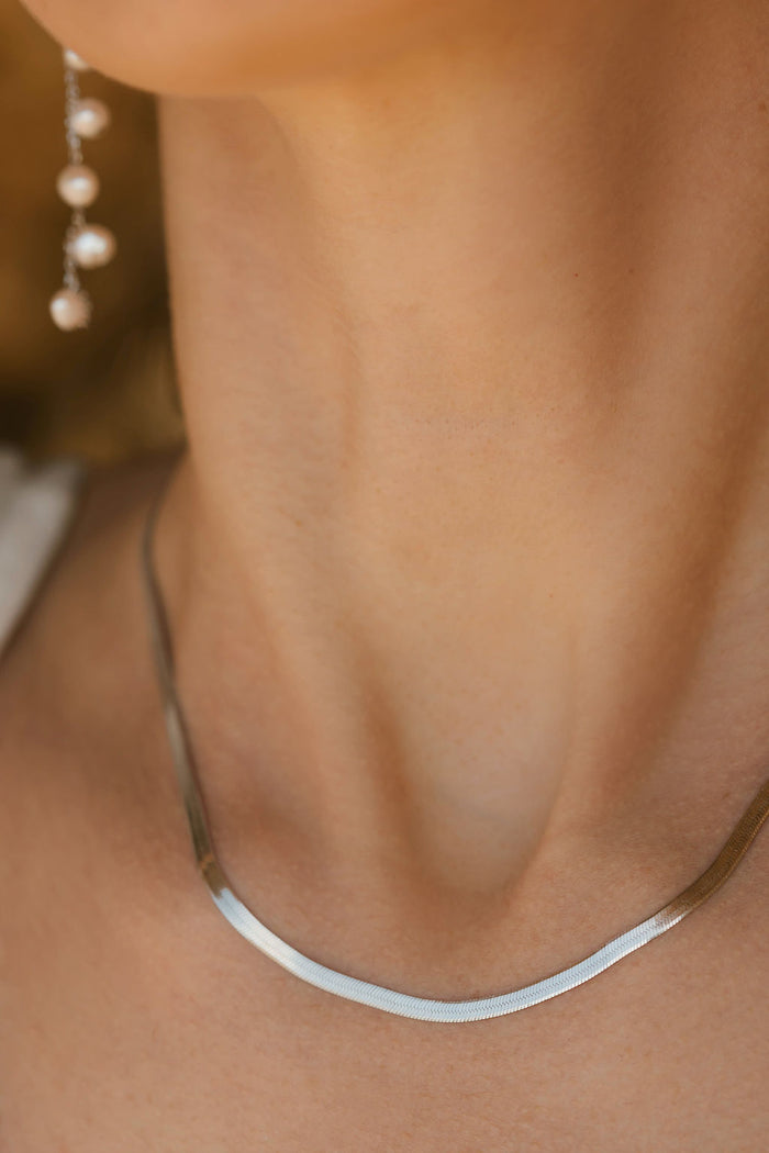 Close-up of a woman's neck wearing LLUME Jewelry's Skinny Hailey Chain Silver necklace. One side features a dangling earring with small pearls. The skin tone is warm against a softly blurred background.