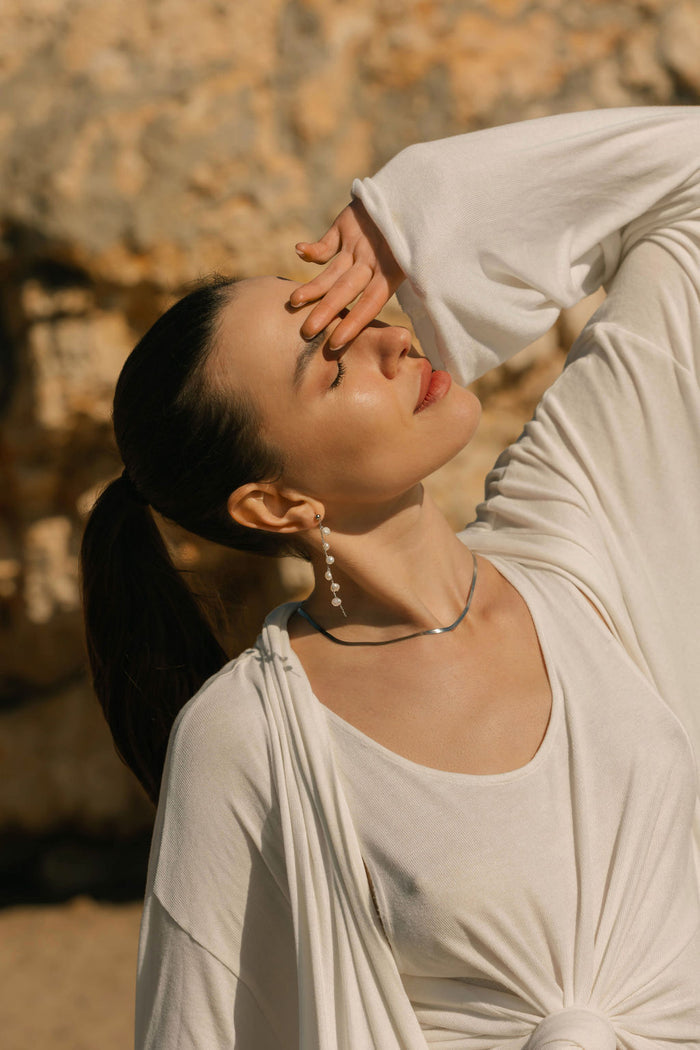 A woman in white stands outdoors, tilting her head back and shielding her eyes from the sun. She wears pearl drop earrings and the LLUME Jewelry Skinny Hailey Chain Silver necklace, with a rocky sunlit background behind her.