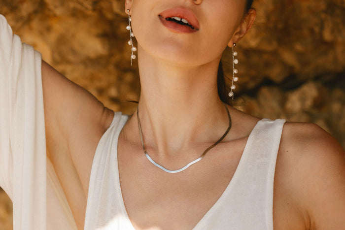 A woman outdoors in a white sleeveless top models the LLUME Jewelry Skinny Hailey Chain Silver necklace, her lips slightly parted and hair pulled back, with dangling pearl earrings and a rocky background behind her.