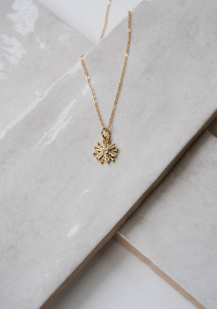 The Snowflake Necklace by LLUME Jewelry features a delicate pendant on a gold filled chain, elegantly displayed on glossy white ceramic tiles—the perfect holiday accessory.