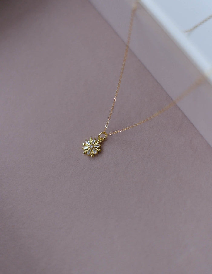 The LLUME Jewelry Snowflake Necklace, featuring a delicate gold filled chain and detailed snowflake pendant, is displayed on a soft mauve background—an ideal piece of holiday jewelry.