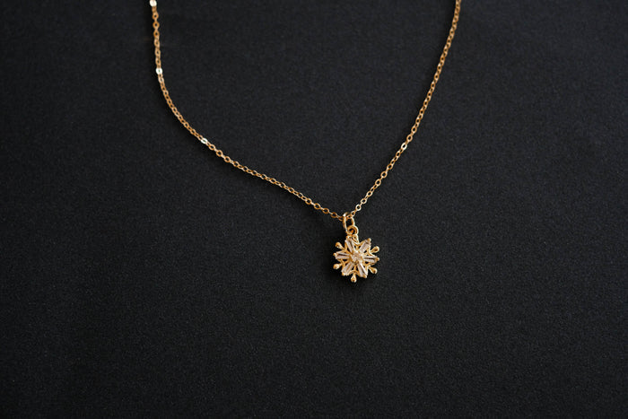 The LLUME Jewelry Snowflake Necklace features a small snowflake-shaped pendant on a gold filled chain. Displayed on a black background, its delicate beauty makes it the perfect holiday jewelry accessory.