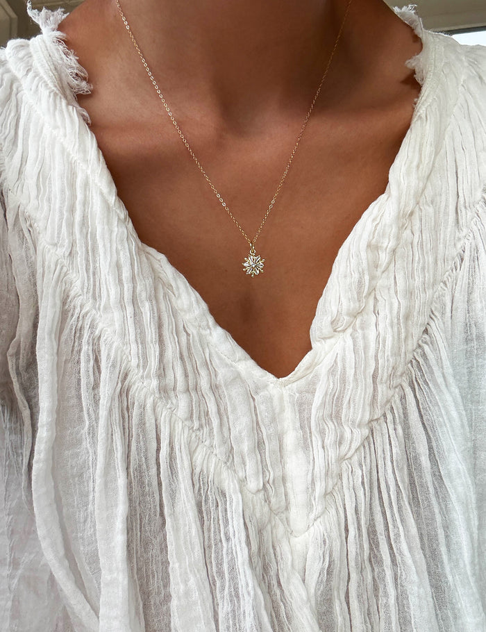 A person wearing a textured white v-neck blouse and the LLUME Jewelry Snowflake Necklace, a delicate gold filled chain with a small starburst pendant, shown from shoulders to chest.