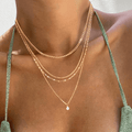 Wearing a green sparkly top, a person showcases LLUME Jewelry's Solstice Set—a gold chain bundle of four layered necklaces with a dainty opal pendant—against a light background.