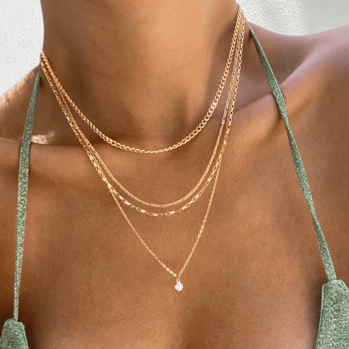 Wearing a green sparkly top, a person showcases LLUME Jewelry's Solstice Set—a gold chain bundle of four layered necklaces with a dainty opal pendant—against a light background.