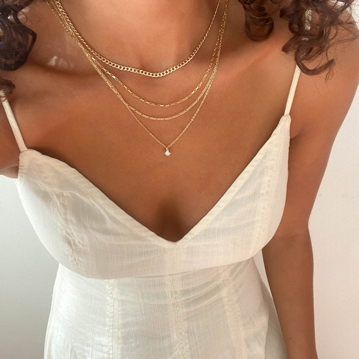 A person wears LLUME Jewelry's Solstice Set—dainty opal pendant and gold necklaces—layered over a white, V-neck, spaghetti strap dress. Only their shoulders to waist show, with curly hair partly visible at the top.