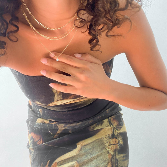 A woman in a strapless printed dress with curly hair leans forward, touching a dainty opal pendant from the Solstice Set by LLUME Jewelry, layered among gold chains. Her light pink nails stand out against a plain white background.