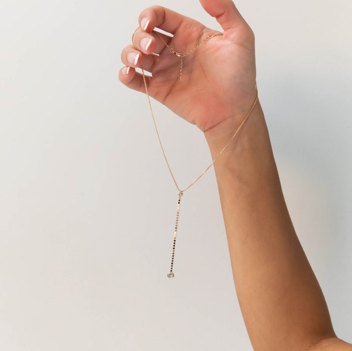 A hand with manicured nails displays the Sophia Necklace by LLUME Jewelry, showcasing a long, vertical 18k gold filled pendant accented with small dark beads against a simple light background.