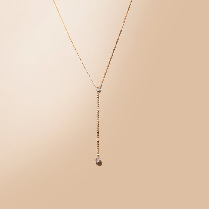 The Sophia Necklace by LLUME Jewelry is an 18k gold-filled necklace with a fine chain, vertical drop, small stones along the drop, and a larger stone at the end, shown on a beige background.