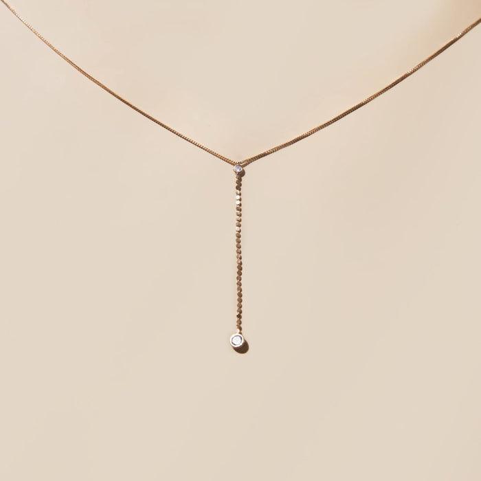 The Sophia Necklace by LLUME Jewelry showcases a delicate 18k gold filled chain with a vertical strand ending in a petite round gemstone, set against a neutral beige backdrop.