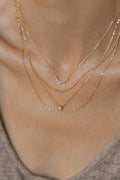 A close-up shows a person wearing the Stillness Capsule Necklace Set by LLUME Jewelry: three delicate gold layered necklaces with a crescent, round gem, and plain chain, styled over a beige top.