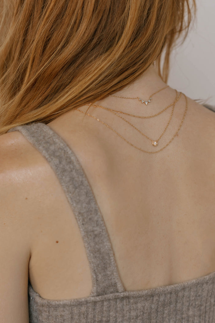 A close-up shows a woman with long, light brown hair wearing LLUME Jewelry's Stillness Capsule Necklace Set—three delicate layered necklaces—paired with a grey, ribbed, sleeveless top.