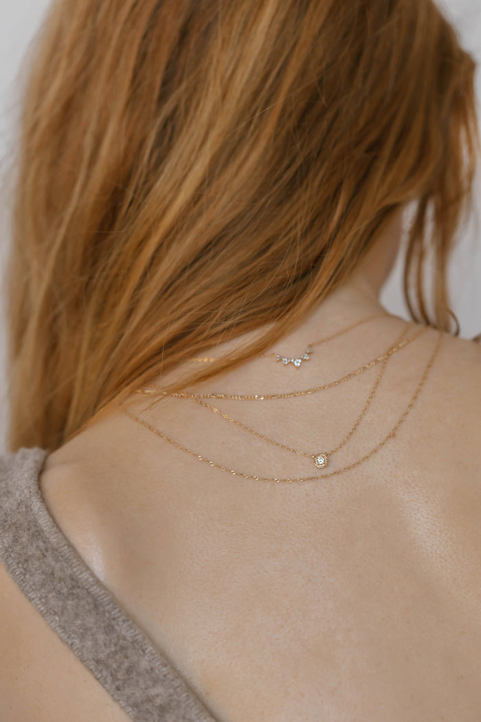 A woman with long auburn hair, seen from behind, wears the Stillness Capsule Necklace Set by LLUME Jewelry—delicate gold pieces layered elegantly over a taupe open-back knit top, draping softly along her upper back and neck.