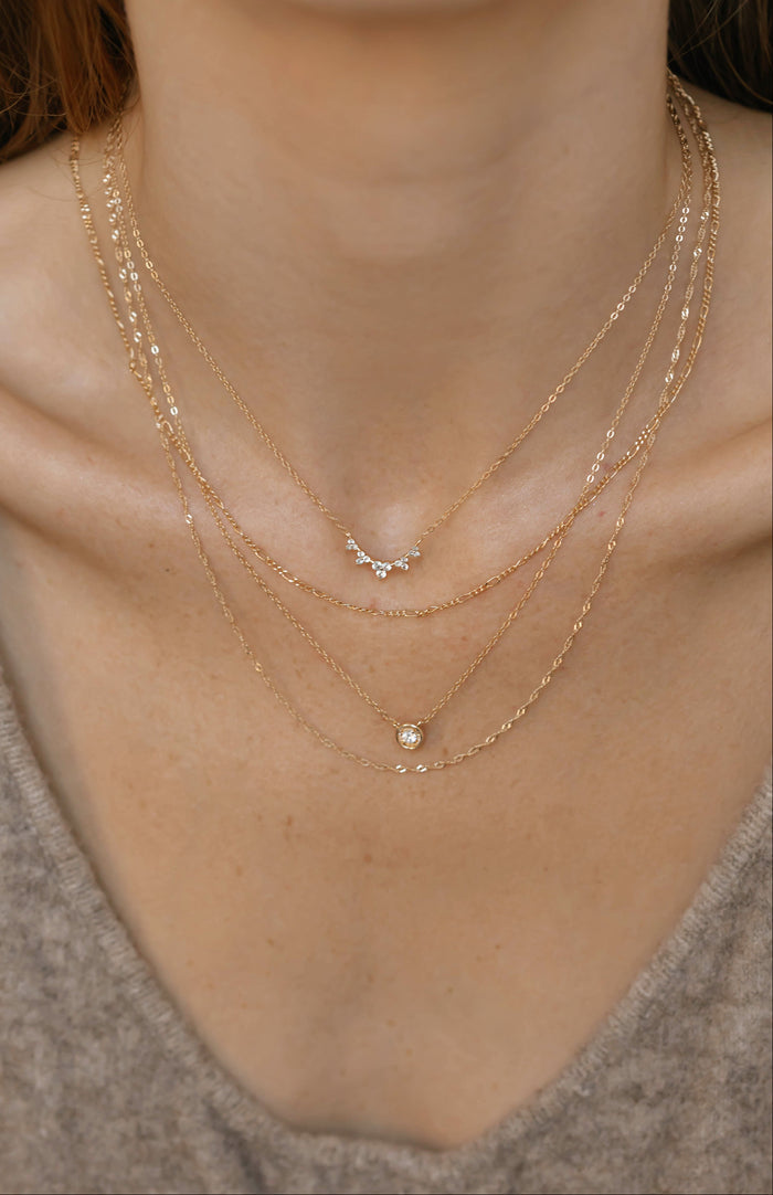 A close-up of a person wearing the LLUME Jewelry Stillness Capsule Necklace Set, featuring three elegant gold chains with delicate pendants, styled over a beige V-neck top.