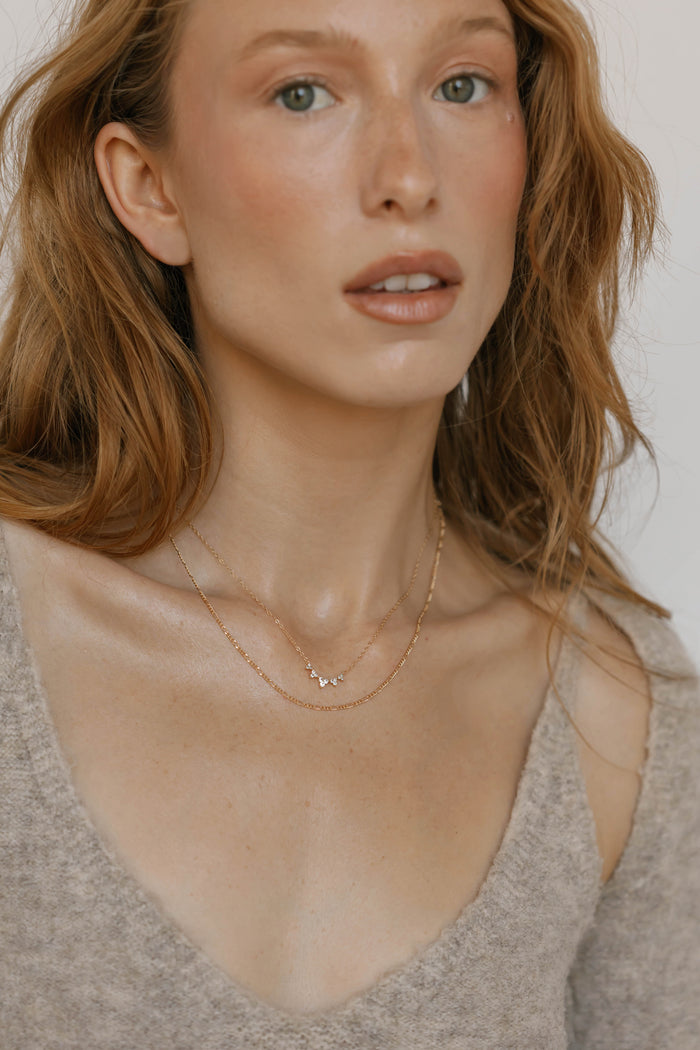 A woman with long wavy light brown hair, fair skin, and minimal makeup wears a beige knit top and LLUME Jewelry’s Stillness Capsule Necklace Set, gazing neutrally just above the camera.