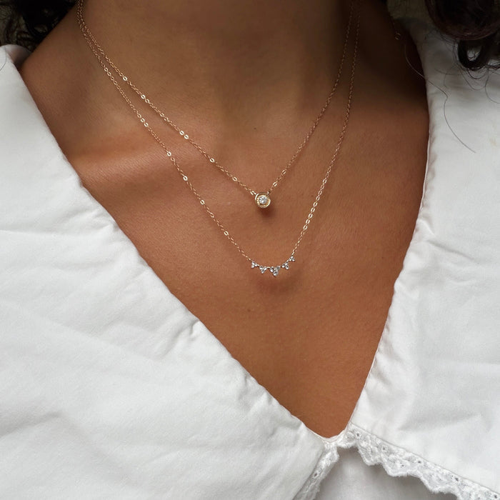 A person wears the LLUME Jewelry Stillness Capsule Necklace Set—layered gold necklaces, one with a round pendant and the other with sparkling stones—styled with a white V-neck blouse for a chic, stacked look.