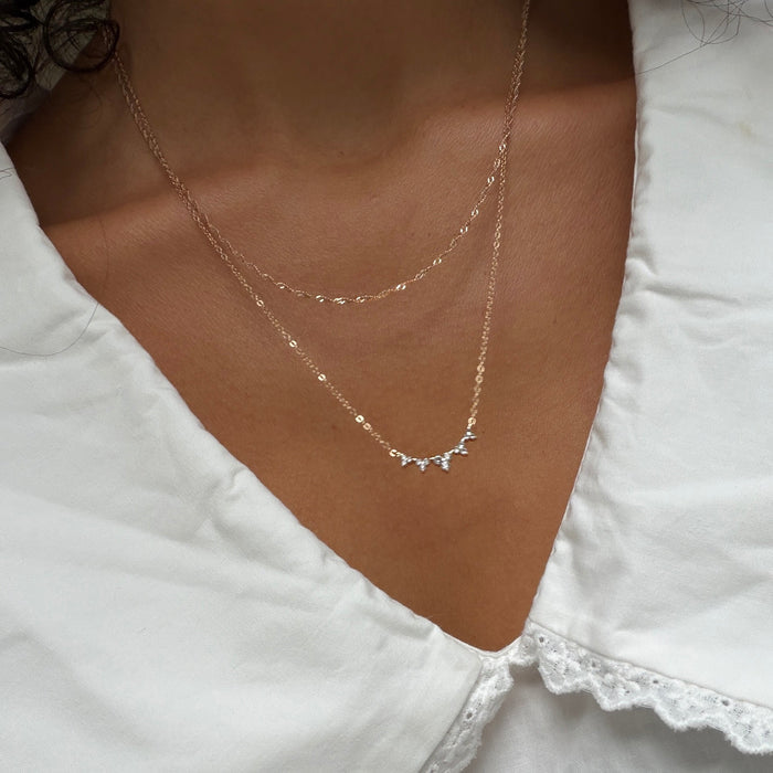 A close-up of a person wearing a white blouse with a scalloped collar, styled with the LLUME Jewelry Stillness Capsule Necklace Set—featuring a gold chain and a charm necklace—for an elegant, layered look.