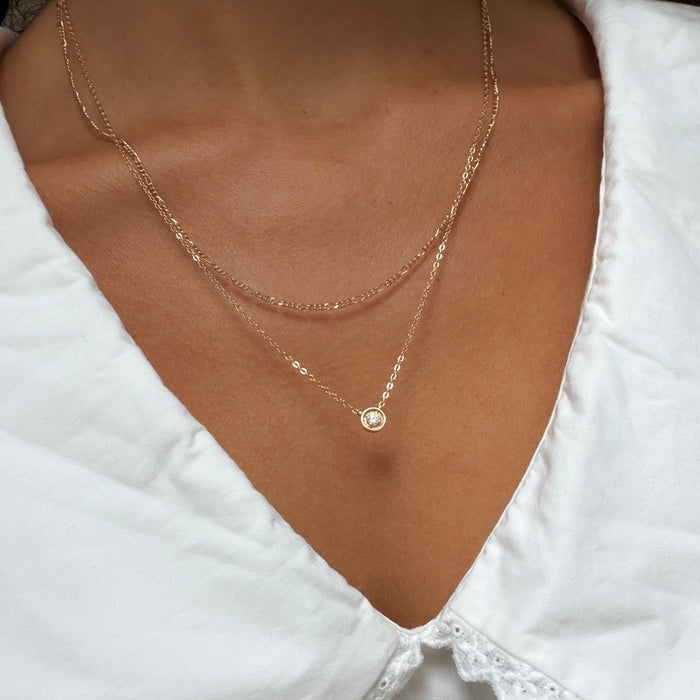 A close-up of a person wearing LLUME Jewelry’s Stillness Capsule Necklace Set—delicate gold pieces, one with a clear gemstone pendant and one simple chain—layered elegantly over a white blouse on their collarbone.