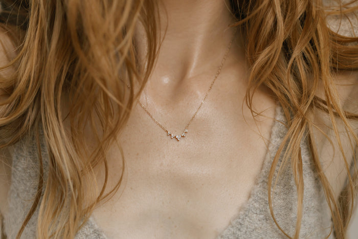 A woman with long, wavy light brown hair wears the Stillness Necklace by LLUME Jewelry—a delicate gold piece with small pendants—paired with a textured gray V-neck top, embodying a mindful, natural style.