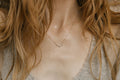 A woman with long, wavy light brown hair wears the Stillness Necklace by LLUME Jewelry—a delicate gold piece with small pendants—paired with a textured gray V-neck top, embodying a mindful, natural style.