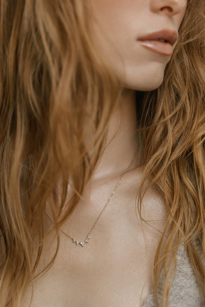 A close-up shows a woman in a grey top wearing LLUME Jewelry’s Stillness Necklace, a delicate gold piece adorned with small gemstones. Her wavy light brown hair frames her face, which is partially out of frame, highlighting the elegant jewelry.