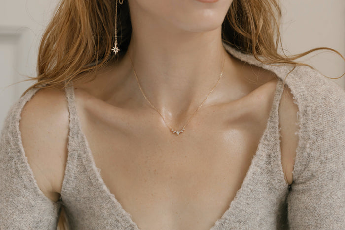 A woman wears a textured beige sweater and gold LLUME Jewelry, featuring the Stillness Necklace with a small star-shaped pendant and matching star drop earrings; her hair is down and her face is partially out of frame.
