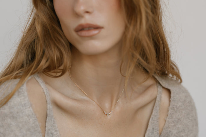 A woman with wavy, light brown hair wears the Stillness Necklace by LLUME Jewelry—a delicate piece featuring small charms. She pairs it with a light gray textured top against a soft, neutral background; her face is partially visible.