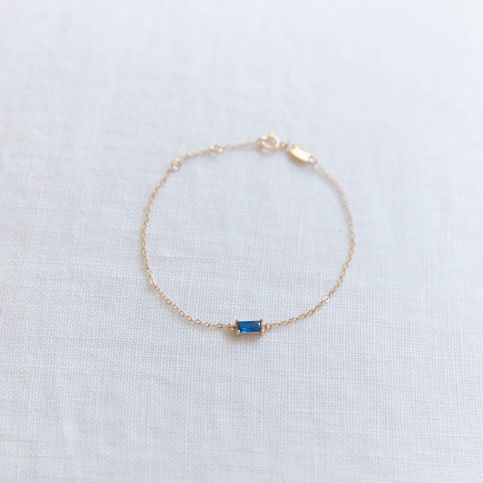 The Taylor Bracelet by LLUME Jewelry features a delicate gold-plated sterling silver chain and a small rectangular blue gemstone centerpiece, beautifully displayed on a light fabric background.