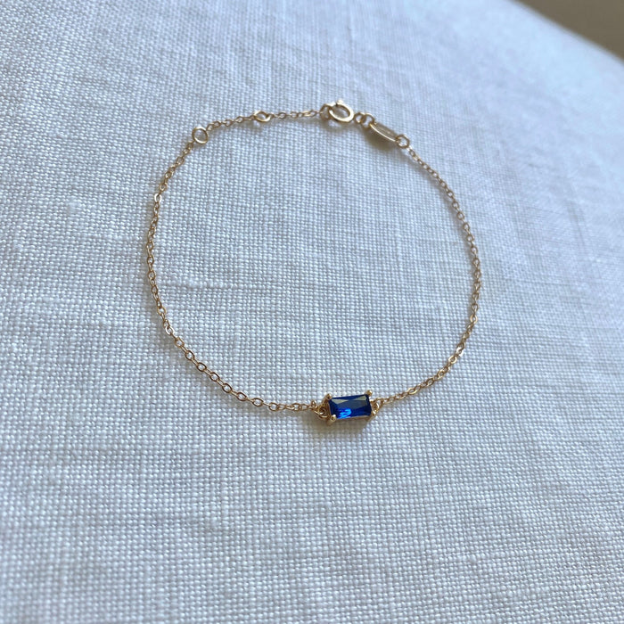 The Taylor Bracelet by LLUME Jewelry features a delicate gold-plated sterling silver chain and a small rectangular blue gemstone centerpiece, shown on textured white fabric.