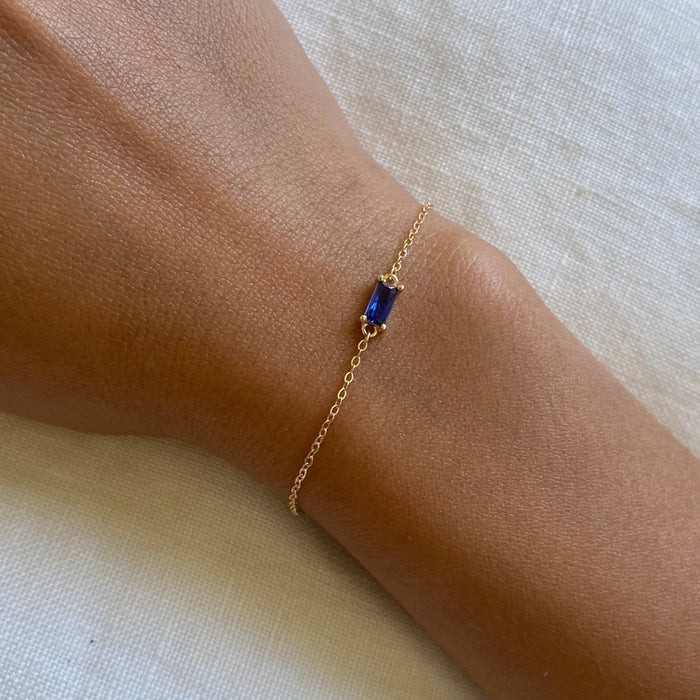 The Taylor Bracelet by LLUME Jewelry features a delicate gold plated sterling silver chain with a small rectangular sapphire blue gemstone, shown worn on a wrist against a light fabric background.