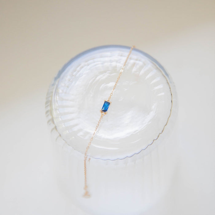 The Taylor Bracelet by LLUME Jewelry features a delicate gold-plated sterling silver chain with a small rectangular blue gemstone pendant, shown draped over clear ribbed glass against a soft, light background.