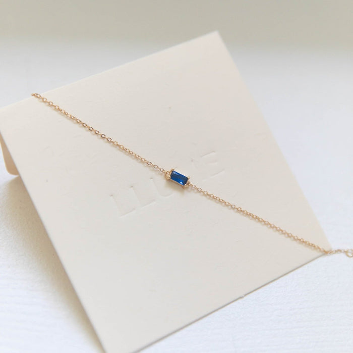 The Taylor Bracelet by LLUME Jewelry features a delicate gold plated sterling silver chain with a small rectangular sapphire blue gemstone, beautifully displayed on a cream-colored card.