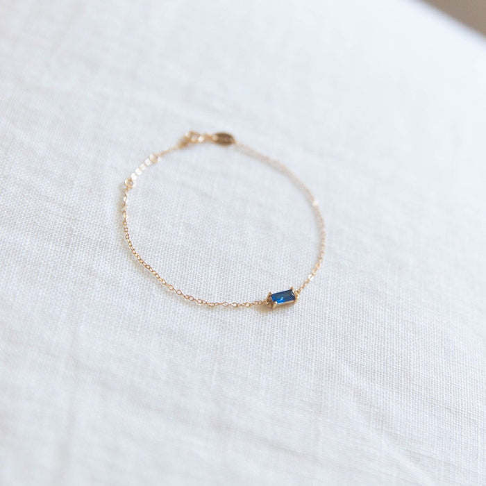 The Taylor Bracelet by LLUME Jewelry, featuring a delicate gold plated sterling silver chain and a small rectangular blue gemstone centerpiece, is displayed on a white textured fabric surface.