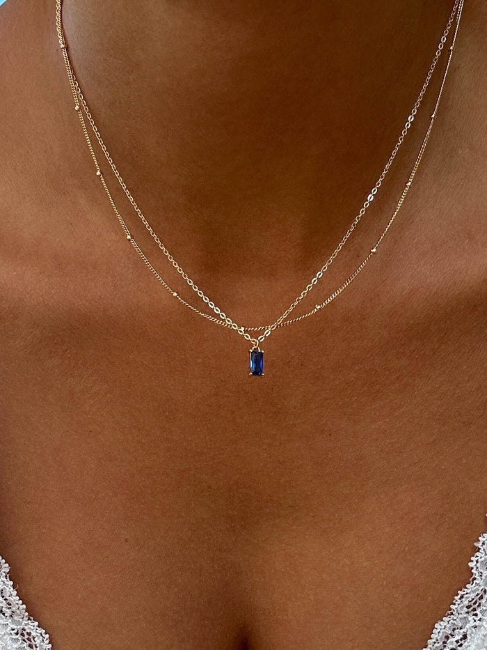 A close-up of a woman's neck wearing the LLUME Jewelry Taylor Choker Set—two layered gold necklaces, one featuring a small blue rectangular pendant. She pairs it with a white lacy top.