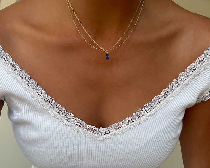 A person wears a white ribbed lace-trimmed V-neck top and the LLUME Jewelry Taylor Choker Set—two delicate gold necklaces, one with a blue pendant. The image is cropped to show the neck, shoulders, and upper chest.