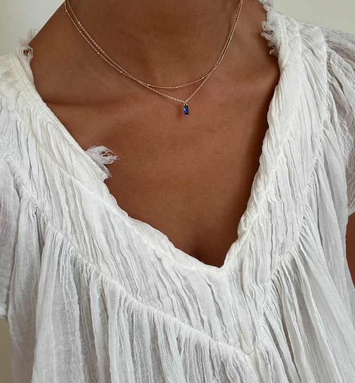 A person wears a crinkled white V-neck blouse and the LLUME Jewelry Taylor Choker Set—layered gold chains with a small blue pendant—highlighting the neckline and jewelry.