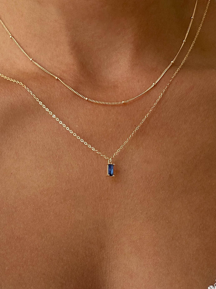 A close-up of a person's neck highlights the LLUME Jewelry Taylor Choker Set, styled with a simple gold chain and a bold blue rectangular gemstone pendant for a chic layered necklace look.
