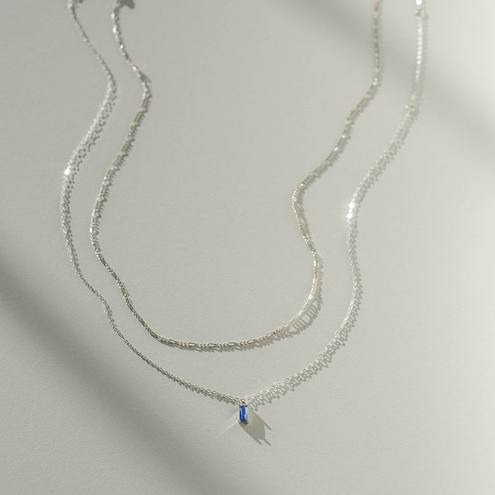 Two delicate silver chain necklaces from the LLUME Jewelry Taylor Daily Set Silver are displayed on a light surface. One is plain, and the other features a small blue rectangular pendant. Both cast gentle shadows in soft light.