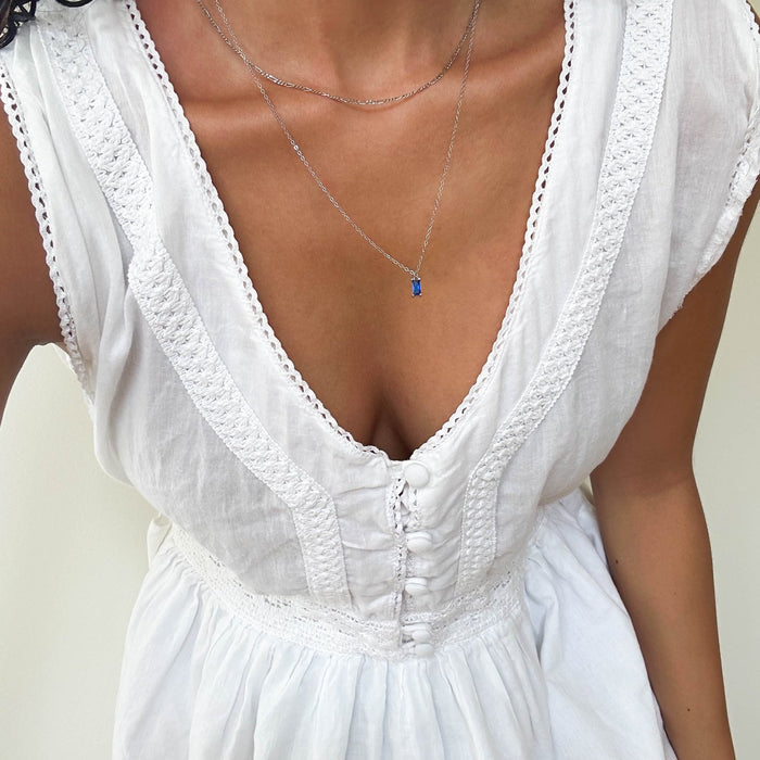 A person in a white, textured v-neck dress with decorative trim and buttons wears the Taylor Daily Set Silver by LLUME Jewelry—a delicate silver necklace set. Only the upper body is visible.
