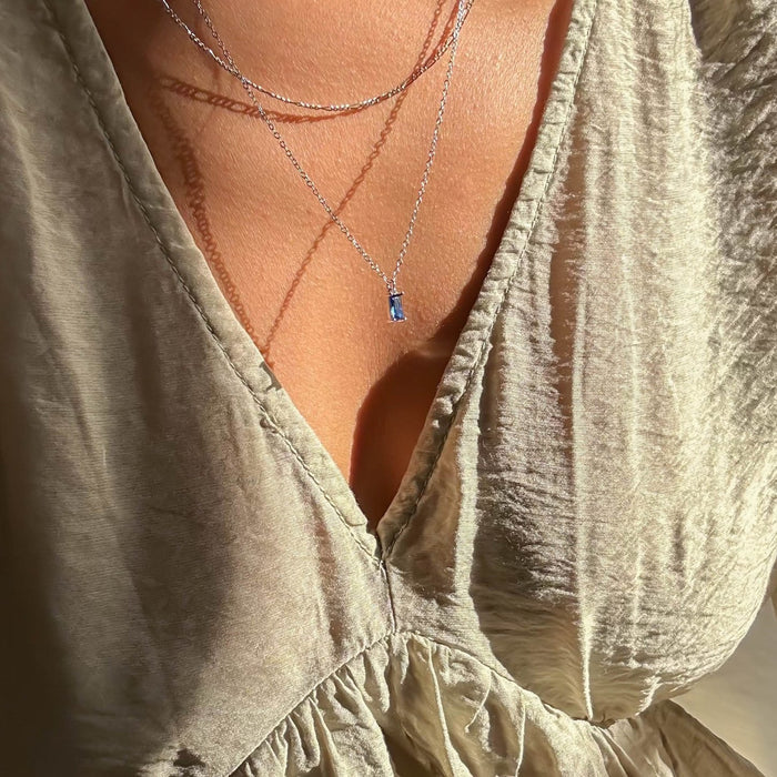 A person wearing a light, shimmery V-neck top and the LLUME Jewelry Taylor Daily Set Silver necklace, featuring a small blue gemstone pendant, shown in warm sunlight.