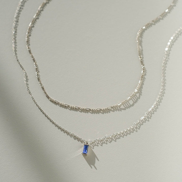 The Taylor Daily Set Silver by LLUME Jewelry features two delicate silver necklaces: a simple chain and the elegant Taylor Necklace with a rectangular blue gemstone pendant, displayed on a light background.