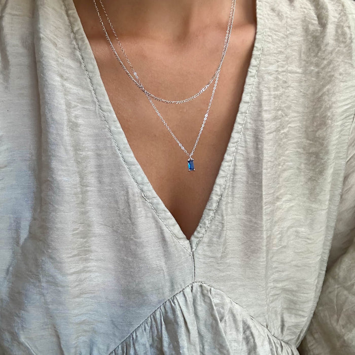 A person wears a light, textured V-neck blouse and the LLUME Jewelry Taylor Daily Set Silver—a delicate silver necklace set with a small rectangular blue pendant—elegantly layered for a refined look.