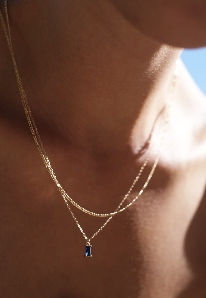 A close-up of a person’s neck wearing the LLUME Jewelry Taylor Daily Set: layered necklaces, including the Taylor Pendant with a small rectangular blue charm. Soft lighting highlights the jewelry against bare skin.