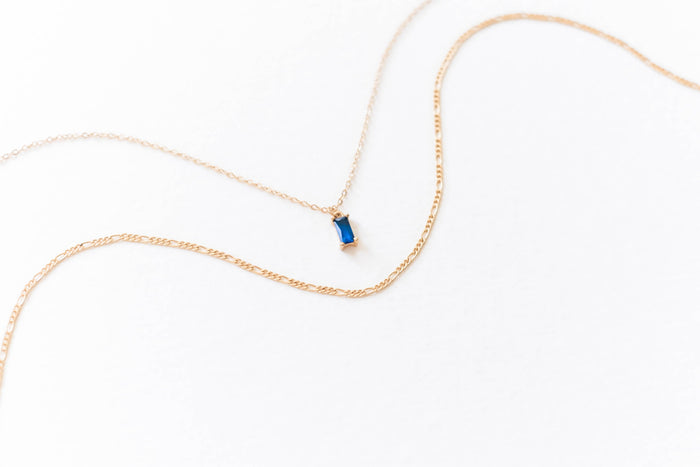 The LLUME Jewelry Taylor Daily Set features a delicate gold pendant necklace with a thin chain and small rectangular blue gemstone, paired with a second slim gold chain—ideal for an elegant layered look.
