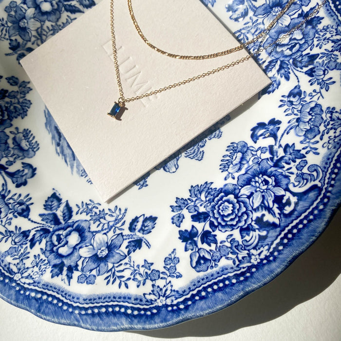 A gold Taylor Daily Set necklace by LLUME Jewelry with a small rectangular blue pendant rests on a white card, displayed on a decorative white and blue floral china plate in bright sunlight.