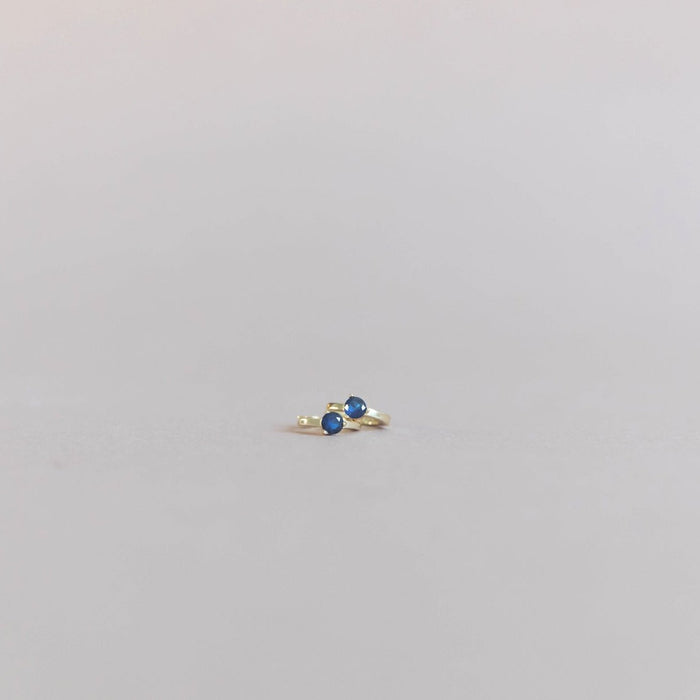 The Taylor Huggies by LLUME Jewelry are small gold-plated earrings with round blue stones, shown on a plain light gray background.