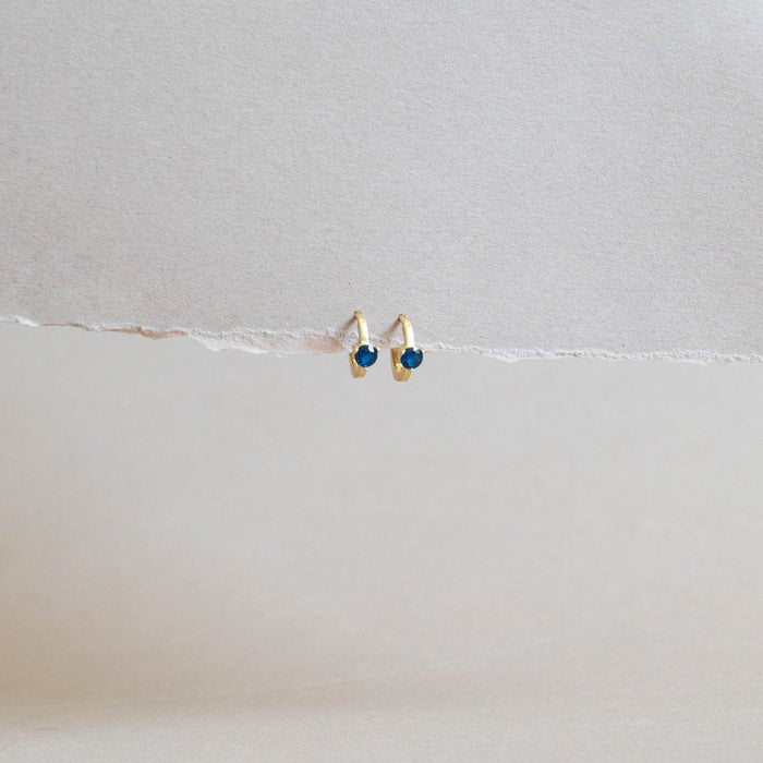 The Taylor Huggies by LLUME Jewelry, featuring small gold plating and deep blue accents, are displayed side by side on textured beige paper with a neutral background.