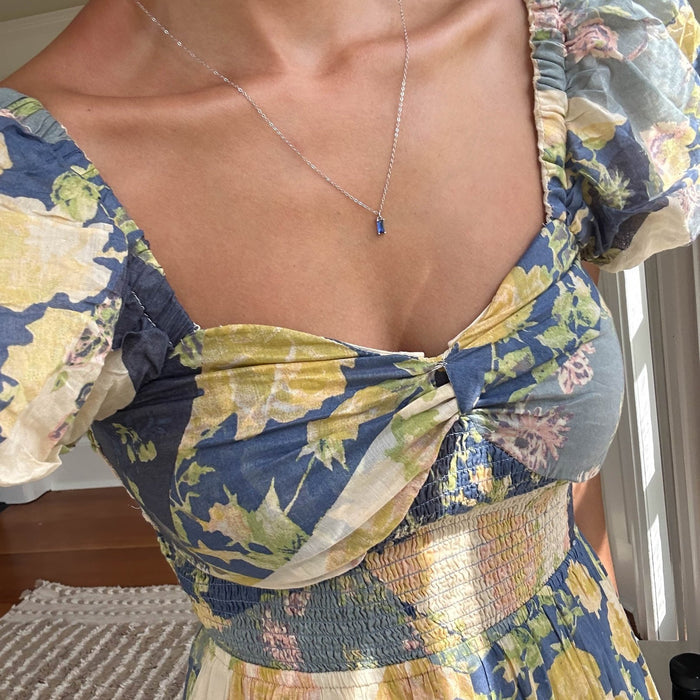Wearing a blue and yellow floral dress with puff sleeves and a ruched bodice, she pairs it with the LLUME Jewelry Taylor Necklace Silver, which features a delicate sapphire blue charm, while standing near a sunlit window.