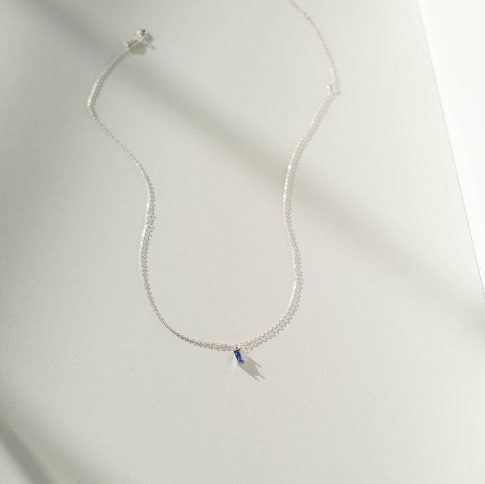 The Taylor Necklace Silver by LLUME Jewelry, featuring a small rectangular blue pendant on a sterling silver chain, is displayed on a white surface with sunlight casting a gentle shadow.
