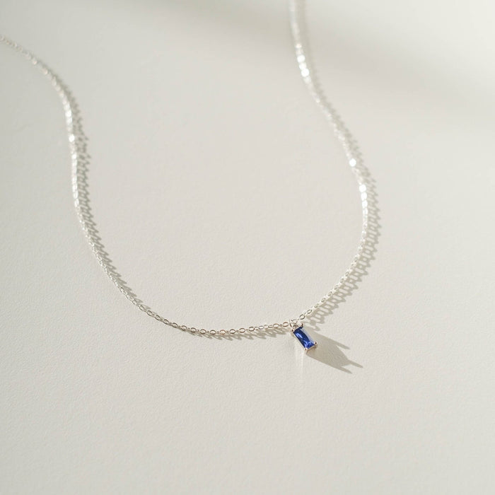 The LLUME Jewelry Taylor Necklace Silver features a delicate sterling silver chain with a small rectangular sapphire blue charm, displayed on a white surface with a soft shadow.
